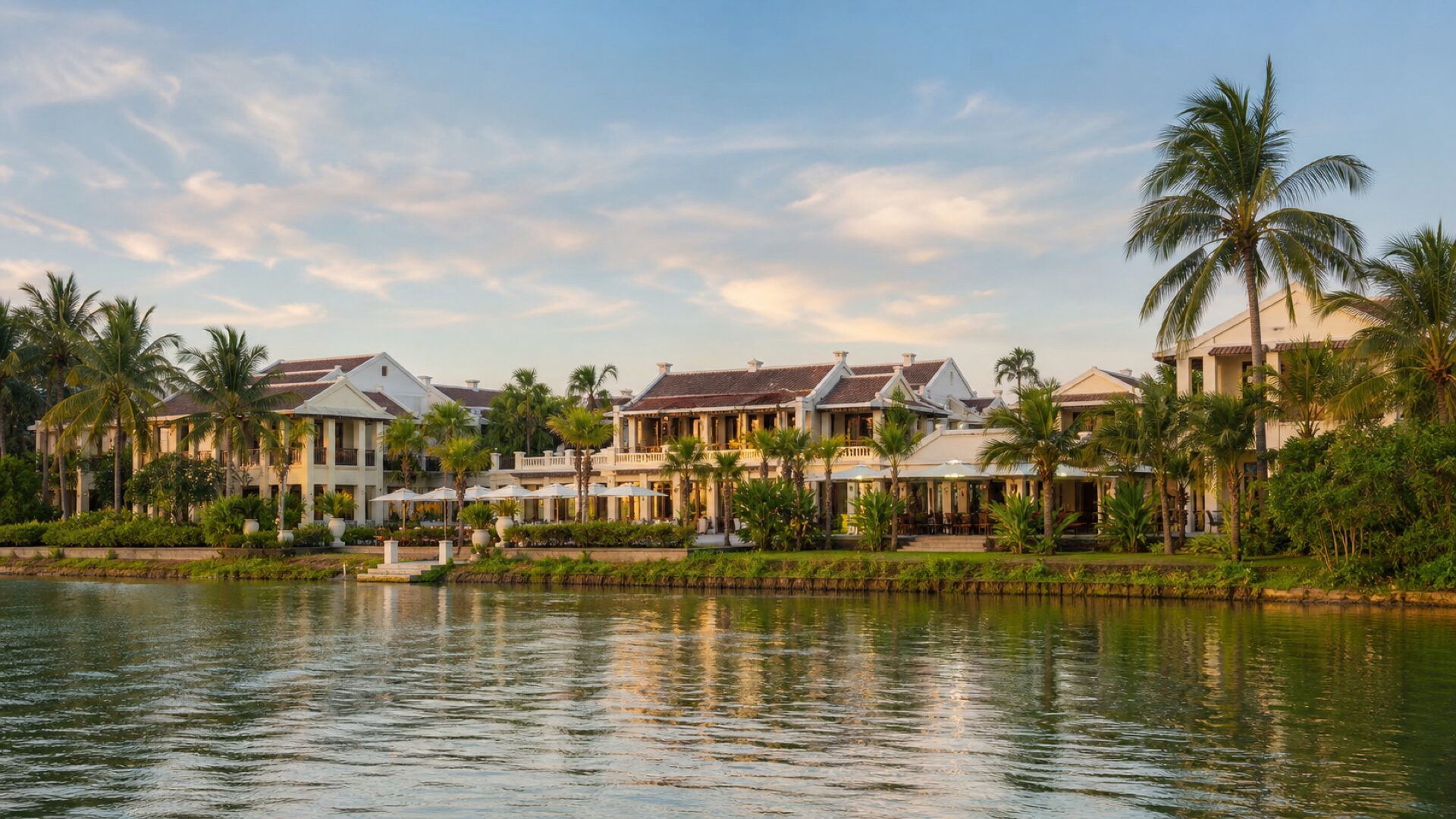 Toàn cảnh Bạch Liên Hội An Resort nhìn từ mép sông Thu Bồn lúc hoàng hôn, tường trắng ngà và hàng dừa hai bên