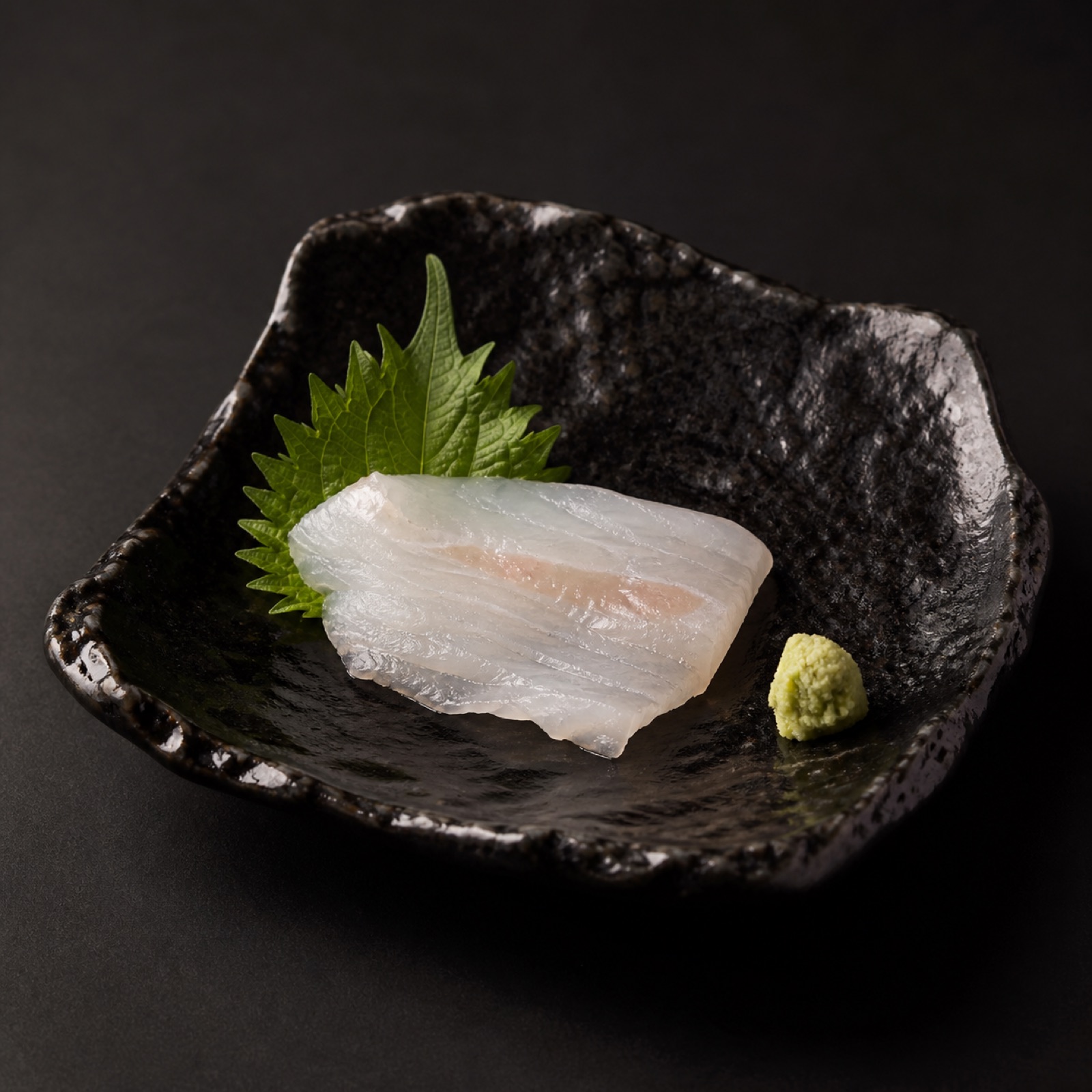 Một món sashimi cá trắng bày trên đĩa gốm đen cùng tía tô và wasabi