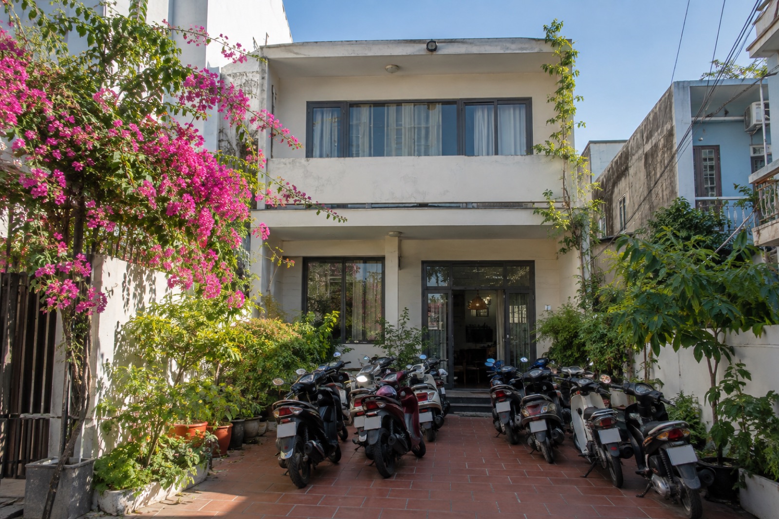 Nhà hai tầng tường vôi trắng, hoa giấy và sân gạch với xe máy đỗ