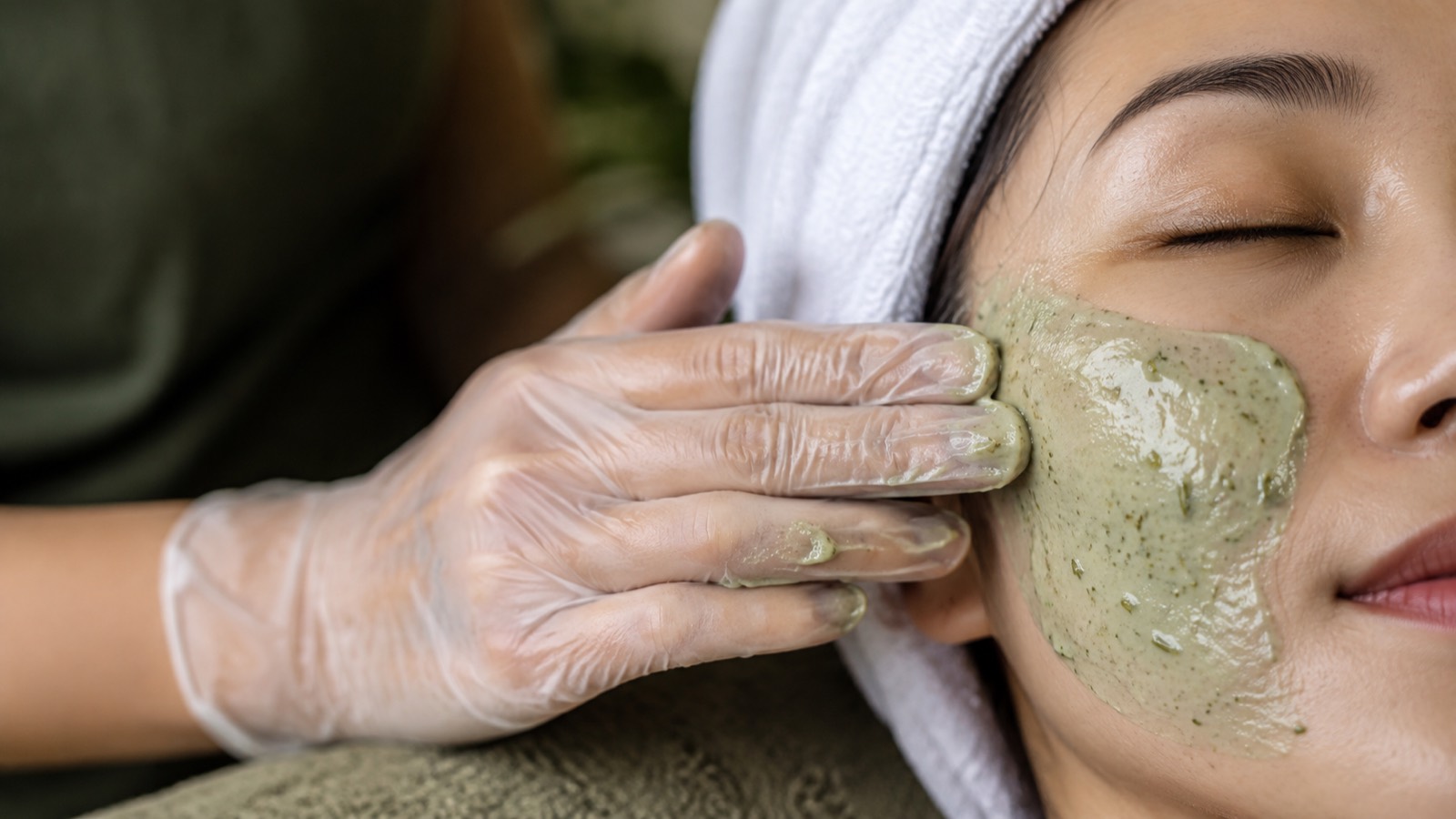 Thoa mặt nạ thảo mộc xanh lên gò má khách nữ đang nhắm mắt, tóc quấn khăn trắng.